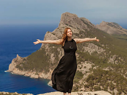 On a rock above the mountains of the Formentor Peninsula in Mallorca, a young woman with red hair stands with her eyes closed, embracing the serene landscape

