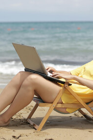 A woman in a yellow shirt sits on a wooden folding chair at the beach, working peacefully by the sea