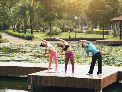An outdoor stretching class takes place by a peaceful lake, with participants practicing Tai Chi near a nearby fountain