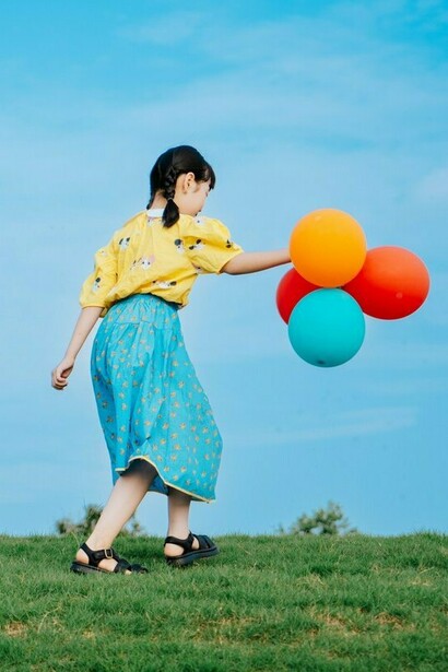 A child extending her hand holding balloons as she moves across the grass, symbolising the instinct to keep something close before it drifts away
