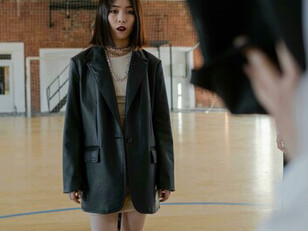 Young girl in an oversized black leather jacket stands on the basketball court as classmates grab her backpack, bullying her
