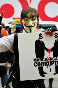 Person wearing a Guy Fawkes mask and holding a sign that reads “March Against Corruption”