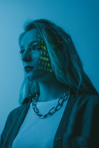 O rosto de uma mulher é iluminado por códigos projetados digitalmente. Estudos apontam que modelos de linguagem ainda associam homens e mulheres a papéis sociais distintos. A inteligência artificial aprende a partir de informações produzidas por sociedades marcadas por desigualdades. Especialistas defendem auditorias e testes mais rigorosos para identificar preconceitos em sistemas de IA