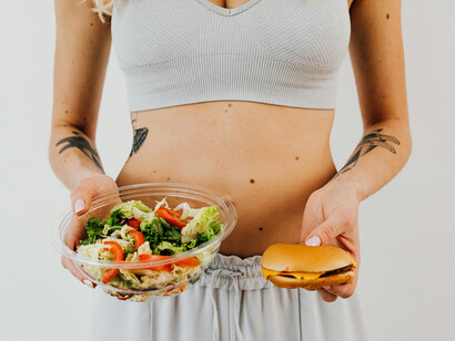 A woman in a white tank top holds a bowl of vegetable salad in one hand and a hamburger in the other