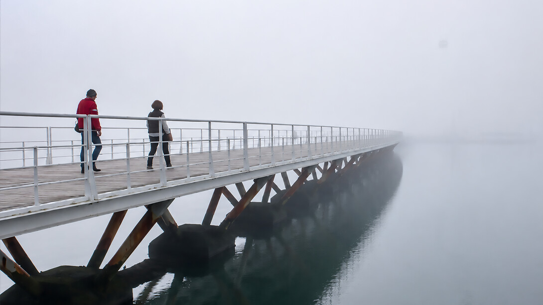 Pedestrians crossing a long wooden bridge over calm water on a misty, fog-covered morning