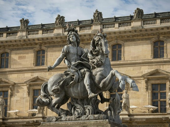 A monumental statue of Louis XIV towering before royal architecture, representing how the execution of a king rarely destroys the structures that sustained his power