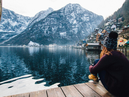 A woman sits on a wooden pier, gazing out at a serene winter landscape of a lake and snow-capped mountains
