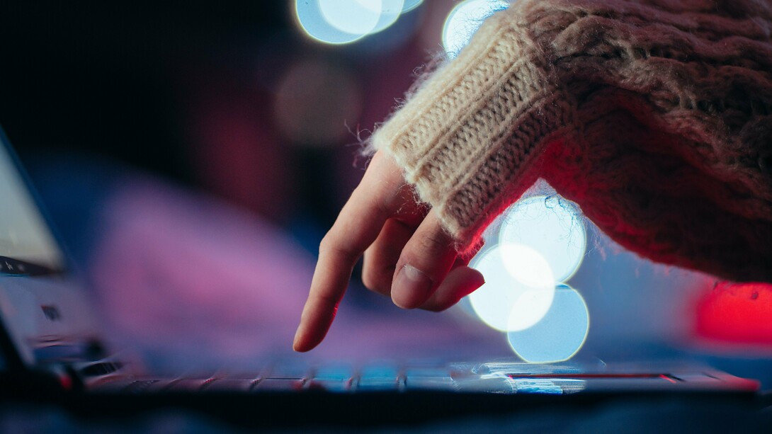 Uma mão pressiona uma tecla de um computador portátil, cercada por pontos de luz desfocados. Um gesto mínimo, quase imperceptível, pode representar o poder de acionar sistemas inteiros — uma metáfora para os riscos de um mundo financeiro cada vez mais dependente de comandos digitais
