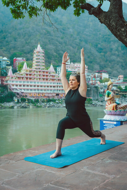 An older woman sits in the lotus position, meditating calmly by the Ganges River in Rishikesh, India — a serene moment of yoga and reflection