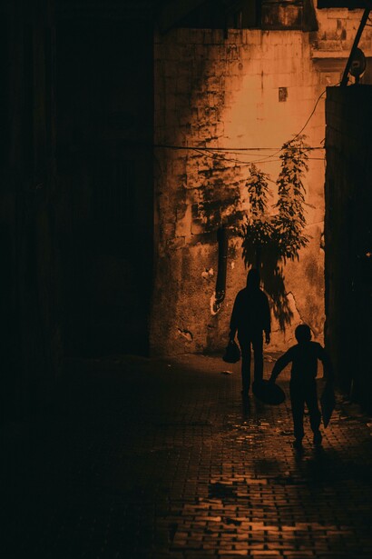Shadowy silhouettes of men linger on a narrow road, while others walk along a dark, foggy pavement
