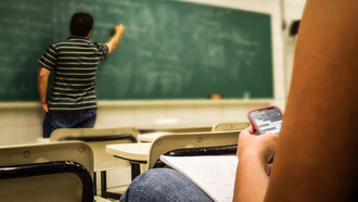 Professor em sala de aula ensinando uma matéria na lousa e a visão do lugar do aluno, que se distrai ao usar o celular