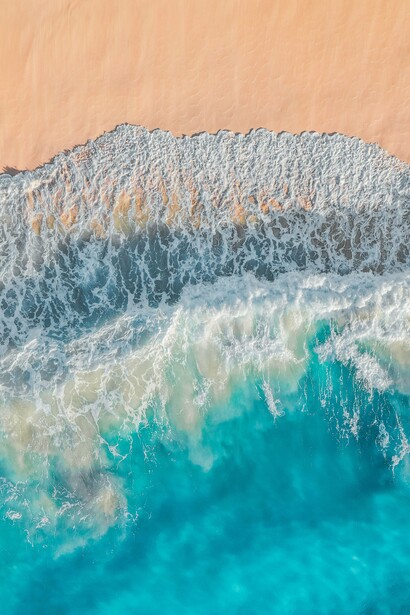 Vista aérea do encontro entre o mar azul-turquesa e a areia, marcado pelo movimento das ondas e pela espuma branca. O contato com a natureza é o primeiro passo para a experiência de desacelerar que a autora associa à viagem