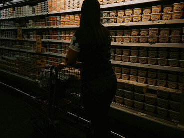 A woman with a shopping cart stands in a softly lit supermarket aisle, surrounded by rows of neatly stocked products