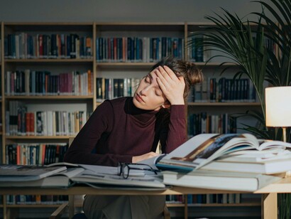 A woman sits at a desk in a library, surrounded by open books, with a weary expression and her head resting on her hand, highlighting the strain of overwork, the lack of work-life balance, and the struggle to achieve a comfortable, fulfilling life