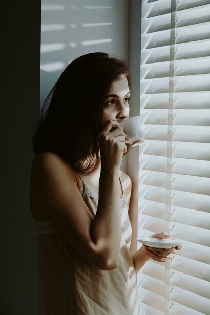 Woman looking out hospital window with sunlight streaming in through the shutters