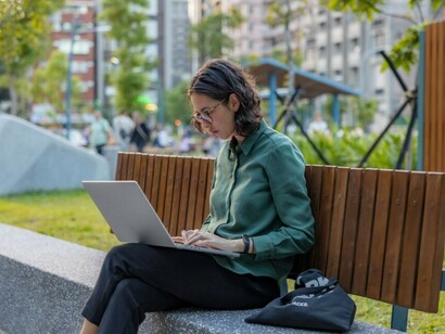 Sunlight filters through the trees as a young woman joins a video call from a park bench — chasing a career dream one click at a time