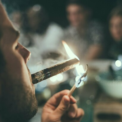 A man lighting a cigar amid a lively setting, an activity not just for the wealthy elite but a timeless pleasure enjoyed by people from all walks of life, united by their appreciation for taste, tradition, and conversation