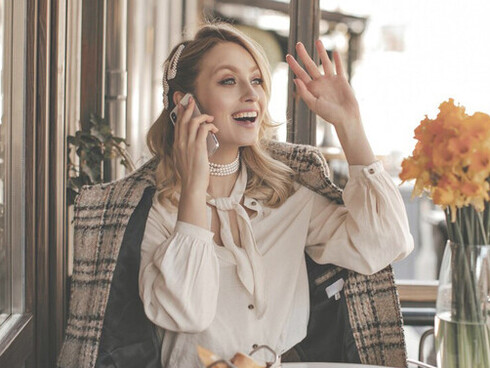 A chic blonde woman in a white blouse adorned with pearl jewelry beams as she sits at a quaint street café, looking off with a joyful expression