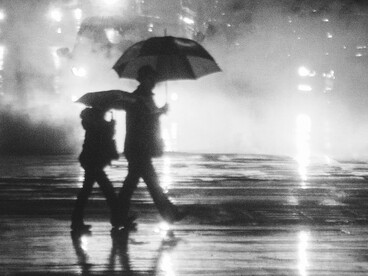 Dos personas caminan bajo la lluvia mientras cruzan una calle iluminada por los reflejos de la ciudad sobre el asfalto mojado