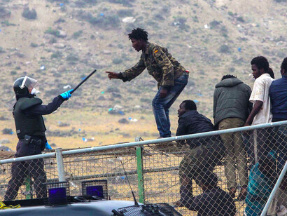 A sub-Saharan migrant agues with a Spanish Guardia Civil officer holding a baton, as they sit on top of a metallic fence that divides Morocco and the Spanish enclave of Melilla, Thursday, May 1, 2014.  (AP Photo/Fernando Garcia)