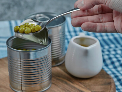 Hand holding a can of boiled green peas on a white table with vegetables and tablecloth