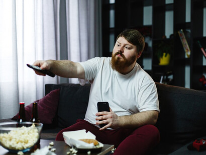 A man lounges on the couch, eating junk food and endlessly switching TV channels, caught in a cycle of inactivity