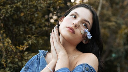 Uma mulher posa ao ar livre com flores delicadas presas ao rosto por um curativo transparente. Às vezes, os pequenos remendos que carregamos contam histórias maiores do que as marcas que procuram esconder. Tal como o botão extra, há gestos discretos que ajudam a manter intacto aquilo que a vida insiste em desafiar. Nem sempre a reparação elimina a marca, mas pode transformá-la em parte da história