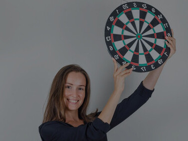 Young woman in black shirt, jeans shorts pointing finger at dartboard and looking hopeful