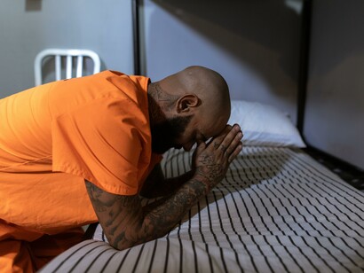 A man in prison leaning on a bed with his hands together