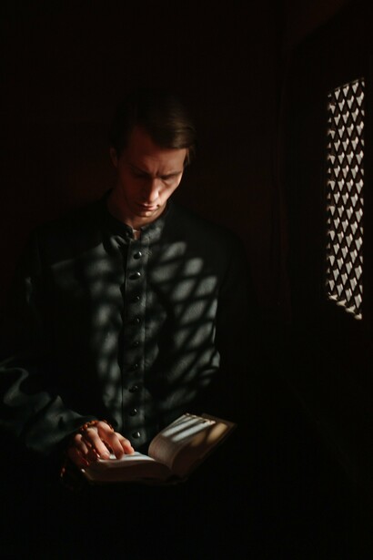 A clergy member holding rosary beads while reading within a confessional, reflecting the need for church leaders to remain anchored in God's Word above financial priorities