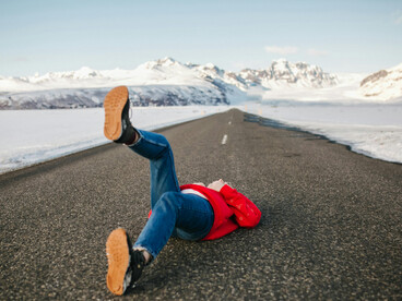 On a quiet Icelandic road, a stylish woman poses while a lone traveler disappears into the distance