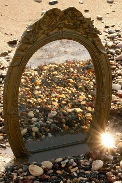 Um espelho de moldura ornamentada repousa entre as pedras à beira-mar, refletindo a paisagem ao redor. Na simbologia de Oxum, o espelho vai além da beleza exterior: convida ao encontro com a própria essência e à descoberta daquilo que permanece em nós, mesmo diante das perdas
