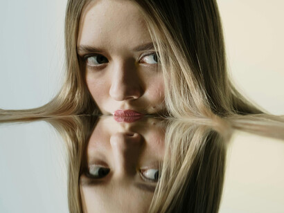 A blonde-haired woman presses her face against a mirror, her expression conveying distress and reflecting inner hypocrisy