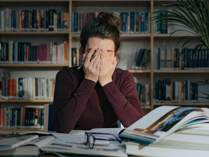 A woman, visibly fatigued, sits in a library with books spread out in front of her, covering her face with her hands, capturing the exhaustion that comes from being overworked and the adverse effects this has on mental health