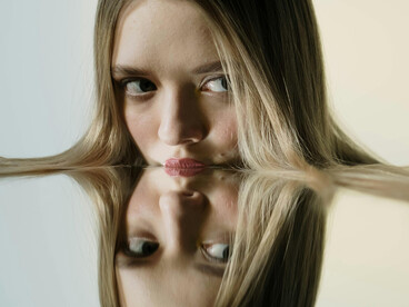 A blonde-haired woman presses her face against a mirror, her expression conveying distress and reflecting inner hypocrisy