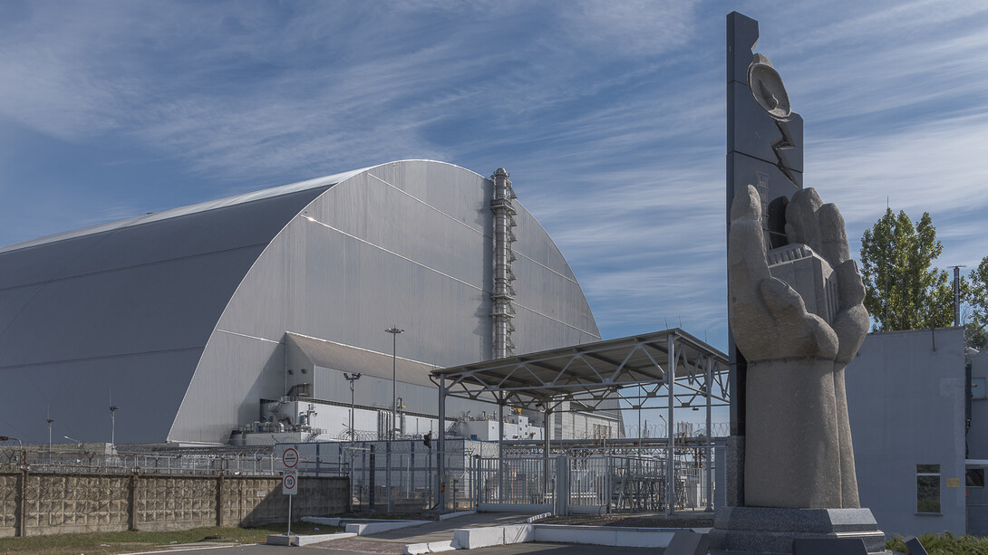 Monumento às vítimas do desastre de Chernobyl em frente ao Novo Confinamento Seguro na Zona de Exclusão de Chernobil, Ucrânia. Os efeitos do desastre de Tchernóbil transcendem fronteiras e gerações, impactando desde a saúde pública local até o cenário energético e político global