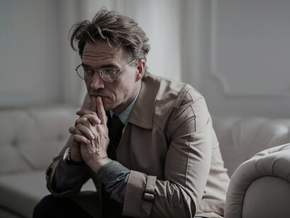 A mature man sits on a couch in a dark living room, deep in thought