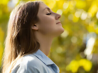 A woman stands in a serene forest, eyes closed and hands on her chest, focusing on deep, calming breaths