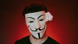 Person wearing a Guy Fawkes mask with a flower tucked behind one ear
