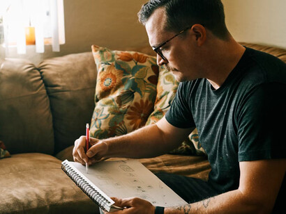 A man in a black crew-neck t-shirt sits on a couch in the early morning light, sketching with quiet focus—lost in a moment of creative inspiration