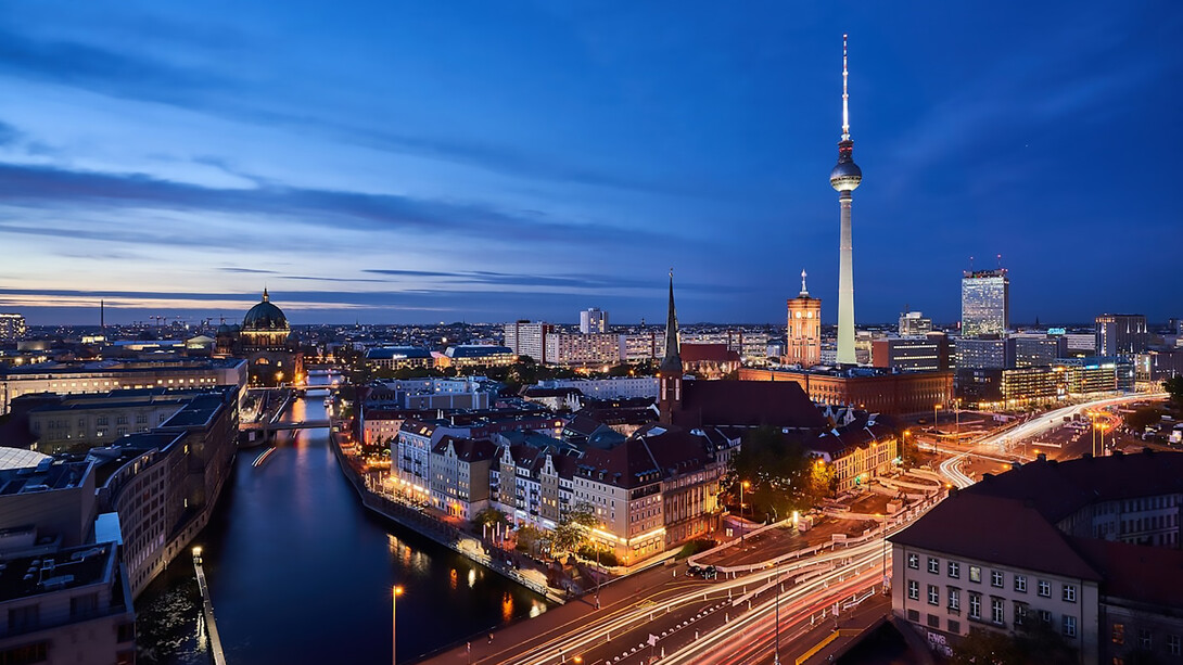 A panoramic scene of Berlin, Germany, reveals the vibrant city at night from a high viewpoint, showcasing the illuminated architecture of Mitte and the serene flow of the Spree River