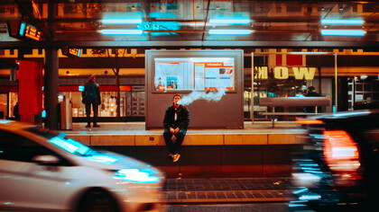 A man sitting on ledge smoking during night time, as neon lights flash by