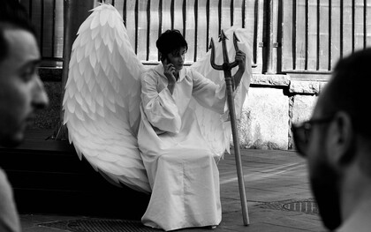 Uma pessoa caracterizada como um anjo, com grandes asas e um tridente nas mãos, sentada enquanto fala ao telefone. A imagem brinca com a dualidade entre aparência e comportamento — justamente a questão que atravessa o artigo: quem somos quando não há ninguém para nos observar? Entre aquilo que mostramos aos outros e aquilo que permanece oculto, o caráter se revela justamente quando não há ninguém para nos observar