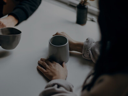 A woman, elegantly dressed in a black long-sleeve shirt, engages in a speed date, cradling a sleek black ceramic mug, while conversing with a gentleman