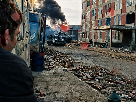 A shattered city street bears the scars of conflict, as smoke rises in the distance and silence settles over the ruins of everyday life