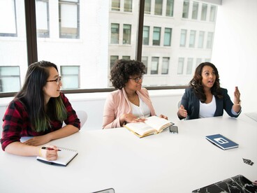 Três profissionais aparecem reunidas diante de uma mesa, em um momento de conversa e atenção compartilhada. O diálogo evidencia o caráter intercambiável da comunicação: quem fala também escuta, e quem escuta pode assumir a palavra