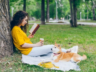 A young woman sitting beneath a tree and reading a book beside her dog, representing the restorative power of quiet leisure and time away from technology