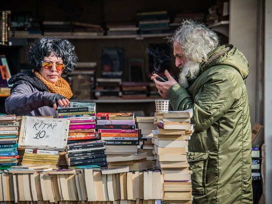 Uma mulher procura um livro; ao seu lado, um homem consulta o celular, ambos cercados por centenas de exemplares. A cena coloca frente a frente duas formas de acesso ao conhecimento e sugere que o desafio do leitor contemporâneo não está apenas em escolher entre o papel e a tela, mas em aprender a transitar entre diferentes possibilidades de leitura. Papel e tecnologia convivem na mesma cena, lembrando que, embora os suportes se transformem, permanece o desejo de ler, descobrir e alcançar novos conhecimentos