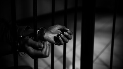 Mãos algemadas se estendem entre as grades de uma prisão, em uma fotografia em preto e branco marcada pelo contraste entre luz e sombra. A imagem evoca o confinamento e a aspiração à liberdade, temas que atravessam o debate sobre a execução penal, a dignidade humana e as possibilidades de reeducação e reinserção social