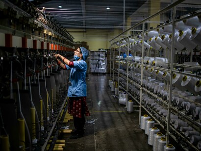 Uma trabalhadora opera máquinas numa fábrica têxtil cercada por fios e bobinas industriais. A modernização da produção trouxe eficiência, mas também expôs gerações de trabalhadores a substâncias químicas perigosas